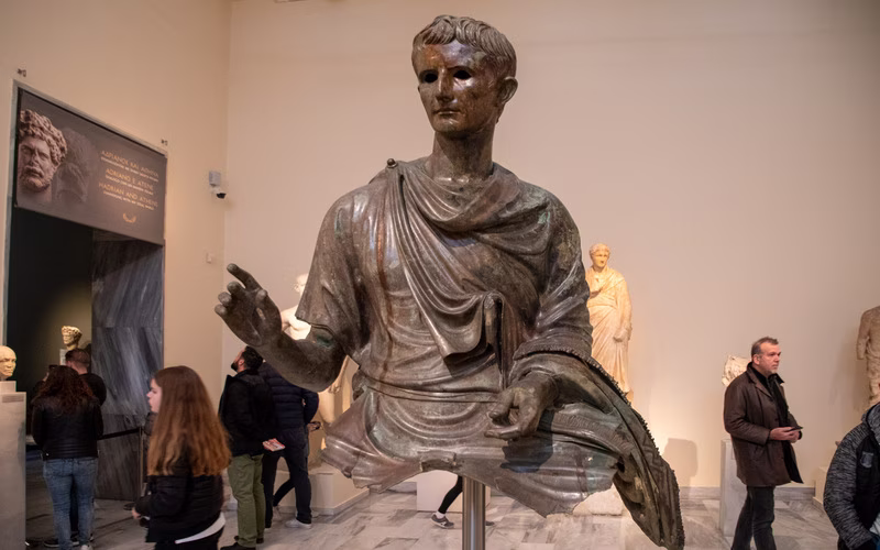 Bronzen beeld van een man in een gewaad in een zaal van het Nationaal Archeologisch Museum