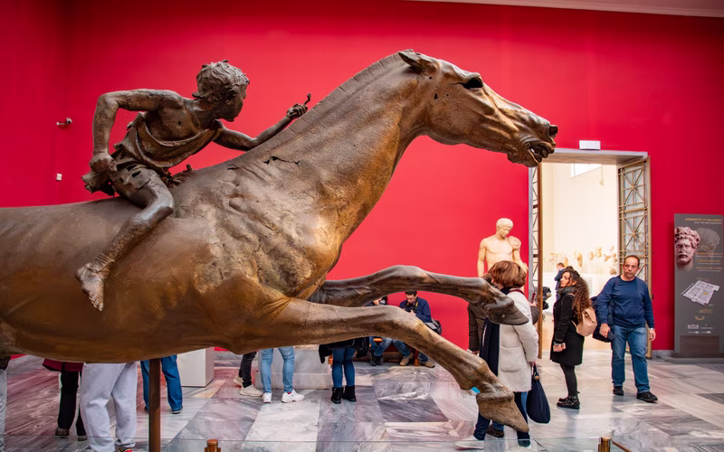 Het bronzen jockeybeeld van Artemision, een jongen op een galopperend paard, tegen een rode wand