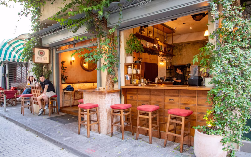 Open cafégevel in Psirri met barkrukken, planten en gasten