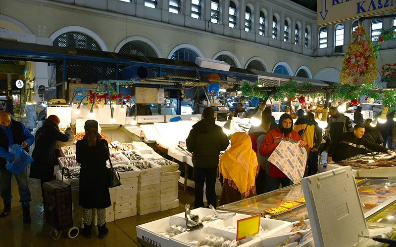 Kopers tussen de viskramen in de centrale markt van Athene, met stapels witte viskisten en de boogramen van de hal erboven