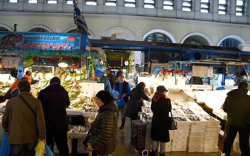 De vishal van de Varvakios-markt met kopers voor de toonbanken, onder de hoge boogramen en met kerstversiering aan de stellingen